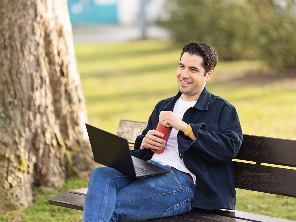 A Henkel employee sits on a bench in a park and works on his laptop.