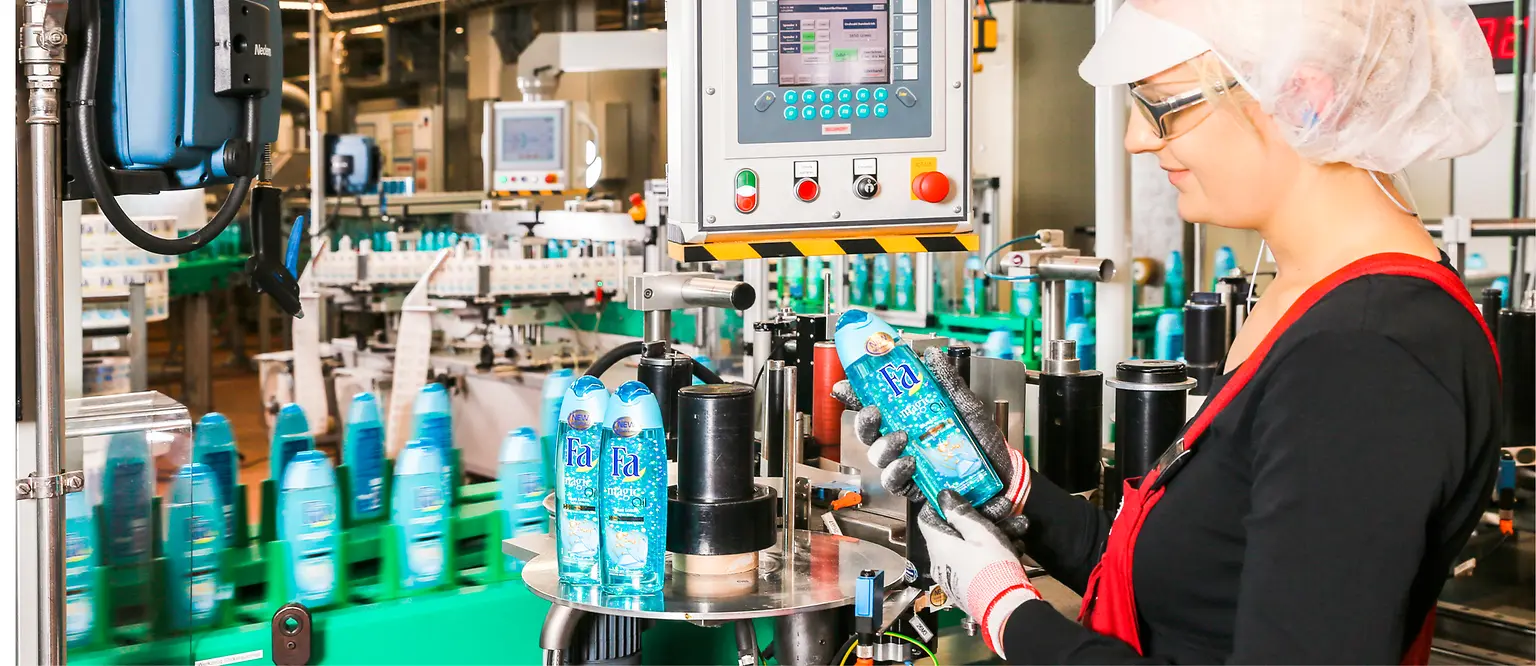 A female employee stands next to the assembly line inspecting a Fa bottle.