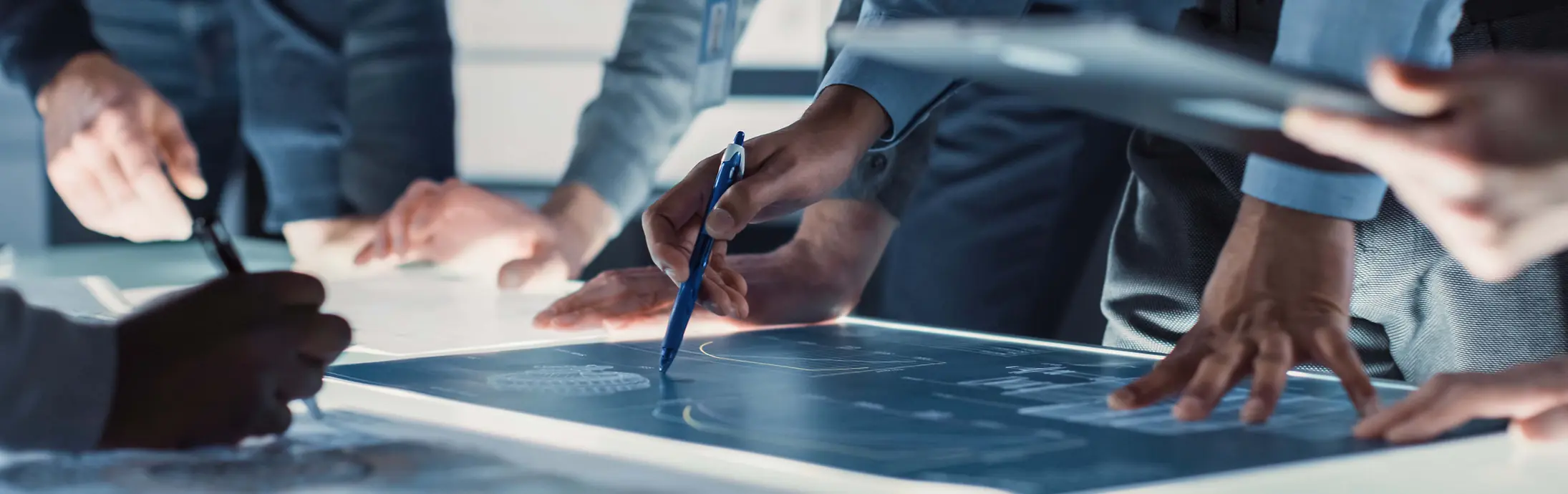 Close-up view of several people gathered around a large illuminated table, reviewing and marking technical blueprints with pens and digital devices.