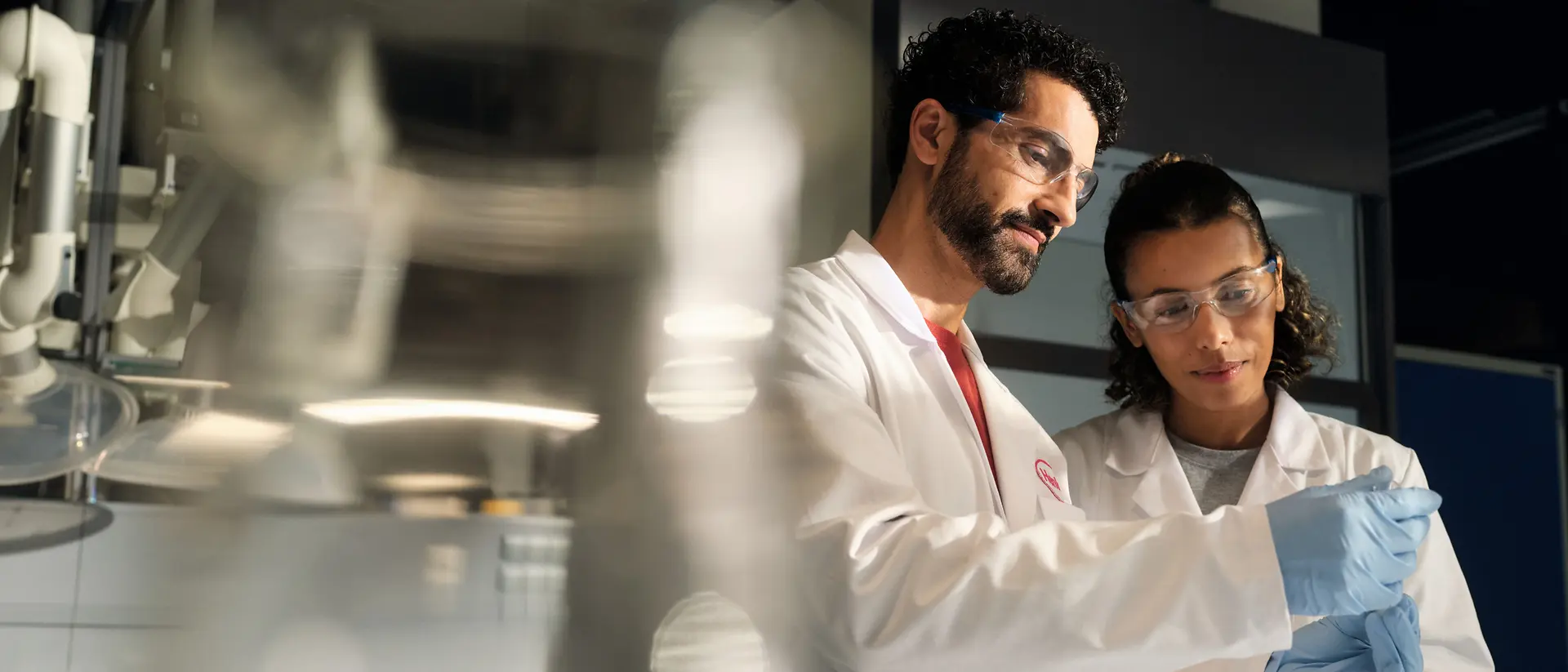 Two scientists wearing white lab coats, safety goggles, and gloves examine a small sample container together in a laboratory setting.