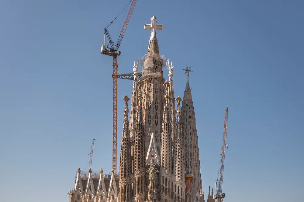 Frontalansicht der Sagrada Família in Barcelona. Im Vordergrund sind die umliegenden Stadtgebäude zu sehen, hinter der Basilika ragen mehrere Baukräne empor. Darüber spannt sich ein klarer blauer Himmel.