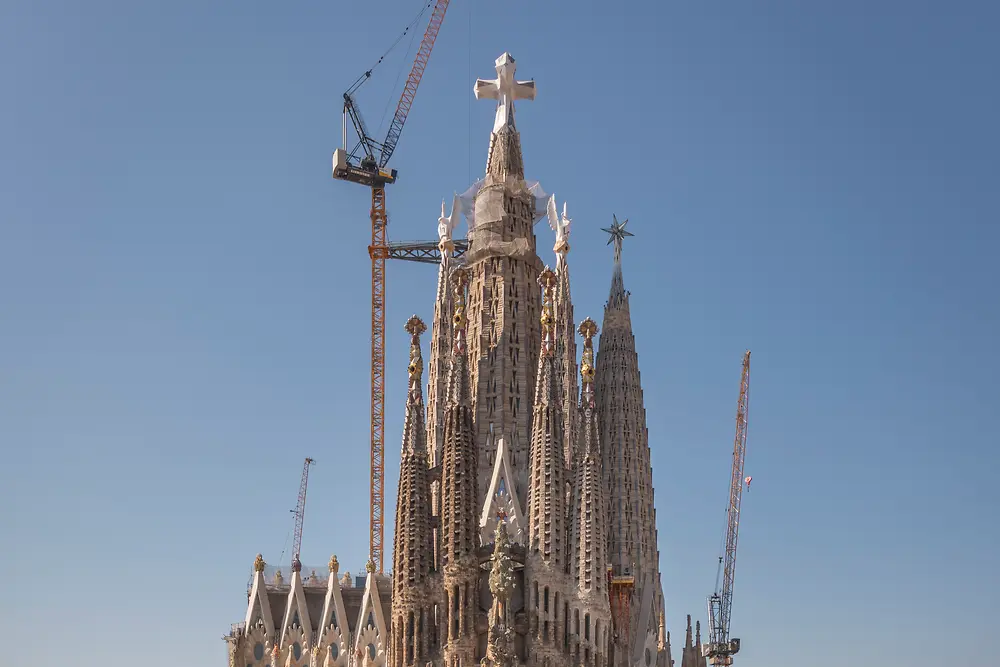 Frontalansicht der Sagrada Família in Barcelona. Im Vordergrund sind die umliegenden Stadtgebäude zu sehen, hinter der Basilika ragen mehrere Baukräne empor. Darüber spannt sich ein klarer blauer Himmel.